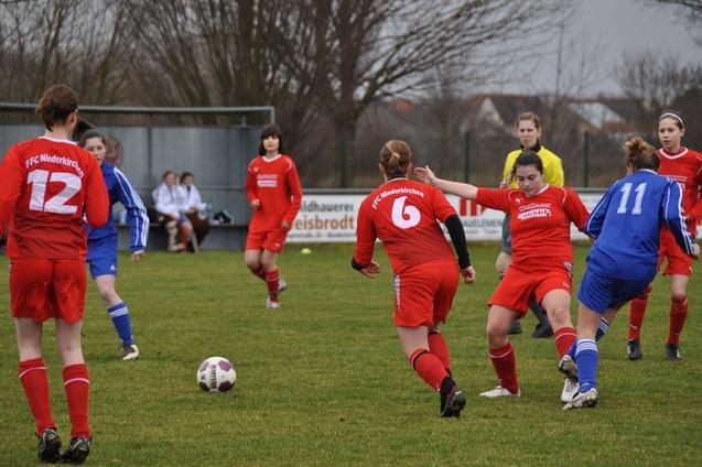 Zwei Gruppen von Fu&szlig;ballspielerinnen in roten und blauen Trikots k&auml;mpfen um den Ball auf einem gr&uuml;nen Spielfeld.