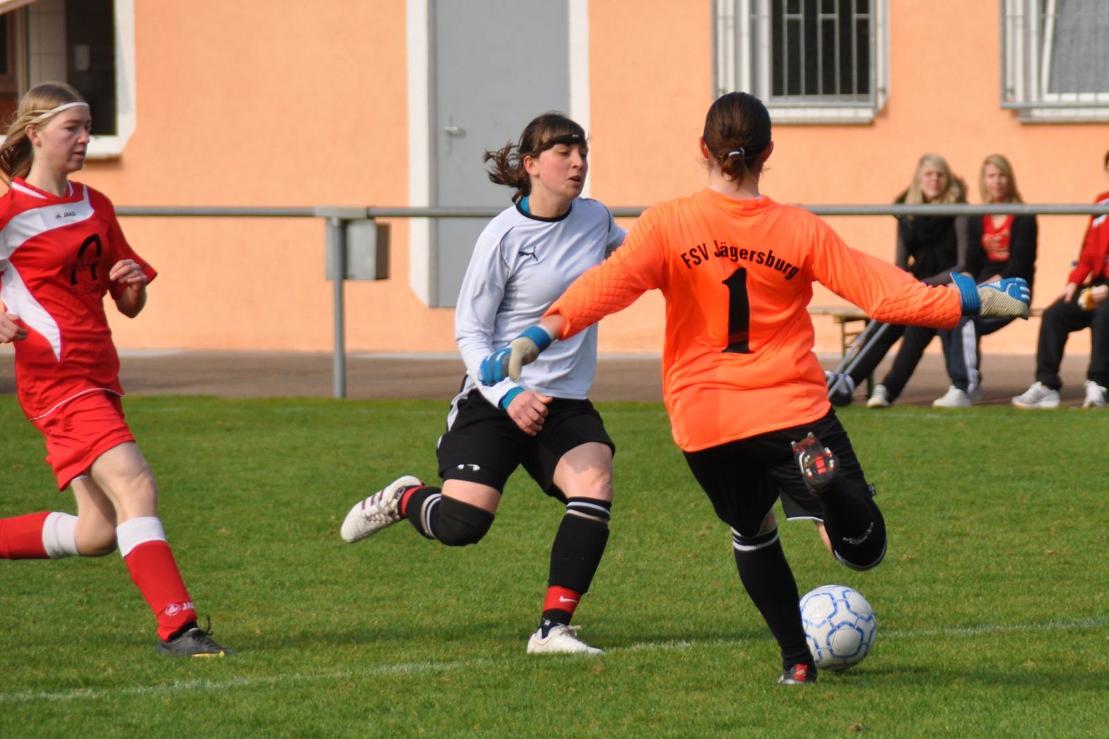 Zwei Fu&szlig;ballspielerinnen im Wettkampf; eine im Tor, die andere greift zum Ball auf dem Rasenplatz.
