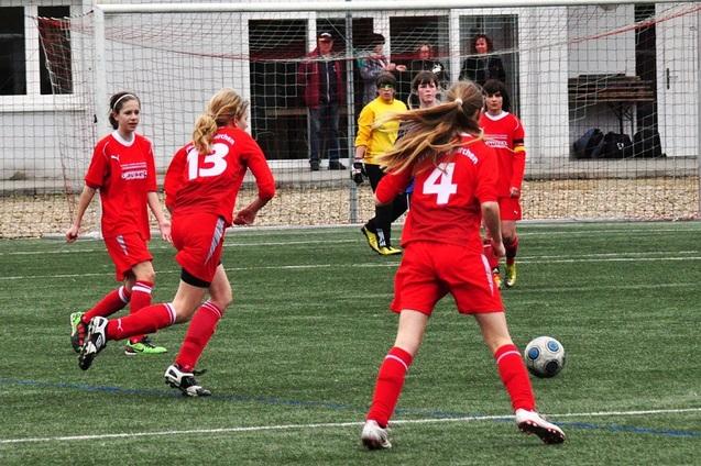 M&auml;dchenfu&szlig;ballspiel auf Kunstrasen, drei Spielerinnen in roten Trikots und eine Torwartin in Gelb, Ball im Spiel.