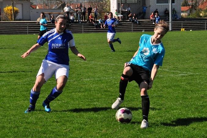 Zwei Fußballspielerinnen kämpfen um den Ball auf einem grünen Spielfeld, im Hintergrund Zuschauer auf der Tribüne.