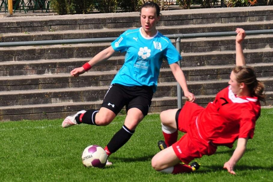 Zwei Fußballspielerinnen in Aktion, die um den Ball auf einem Rasenplatz kämpfen, während eine Spielerin zu Boden fällt.