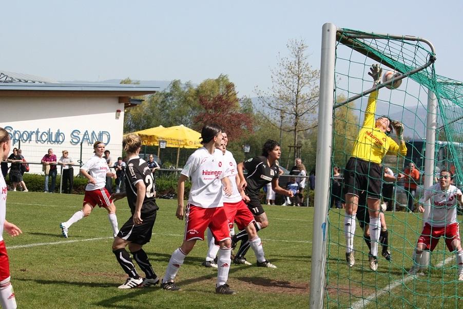Szenenbild eines Fu&szlig;ballspiels: Torh&uuml;ter greift nach dem Ball, w&auml;hrend Spieler in wei&szlig;en und schwarzen Trikots zuschauen.