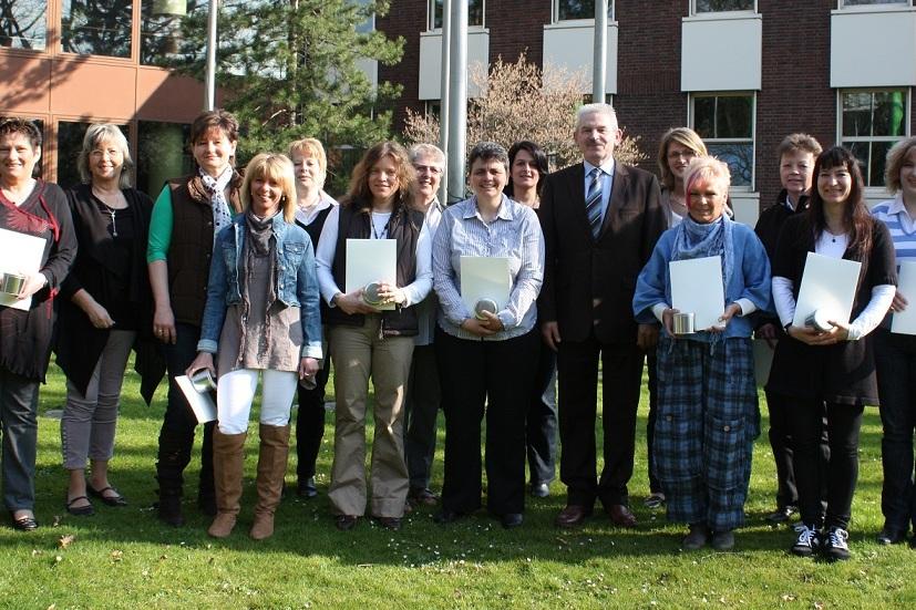 Gruppenfoto von 15 Personen im Freien, einige halten Zertifikate, vor einem Geb&auml;ude mit gr&uuml;ner Rasenfl&auml;che.