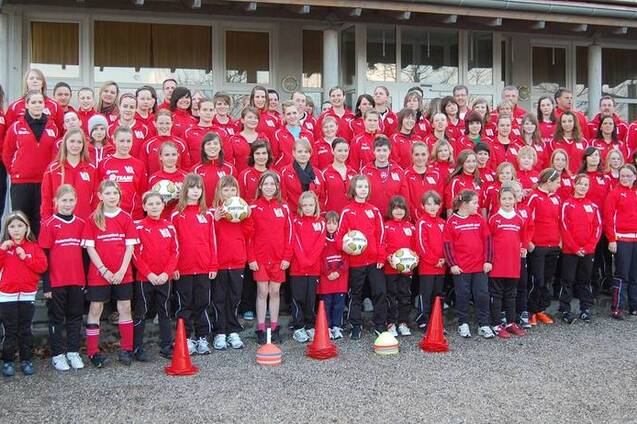 Gruppenfoto von M&auml;dchen und Frauen in roten Trainingsanz&uuml;gen, versammelt vor einem Geb&auml;ude, mit Fu&szlig;b&auml;llen und Trainingsmaterial.