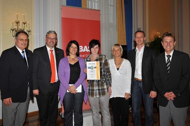 Gruppenfoto von sechs Personen, die eine Urkunde in einem festlichen Raum pr&auml;sentieren, mit einem roten Banner im Hintergrund.