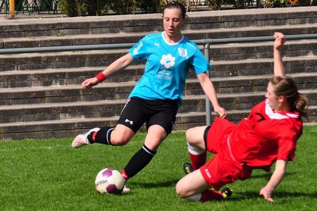 Zwei Fu&szlig;ballspielerinnen in Aktion, die um den Ball auf einem Rasenplatz k&auml;mpfen, w&auml;hrend eine Spielerin zu Boden f&auml;llt.