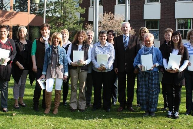 Gruppenfoto von 15 Personen im Freien, einige halten Zertifikate, vor einem Geb&auml;ude mit gr&uuml;ner Rasenfl&auml;che.
