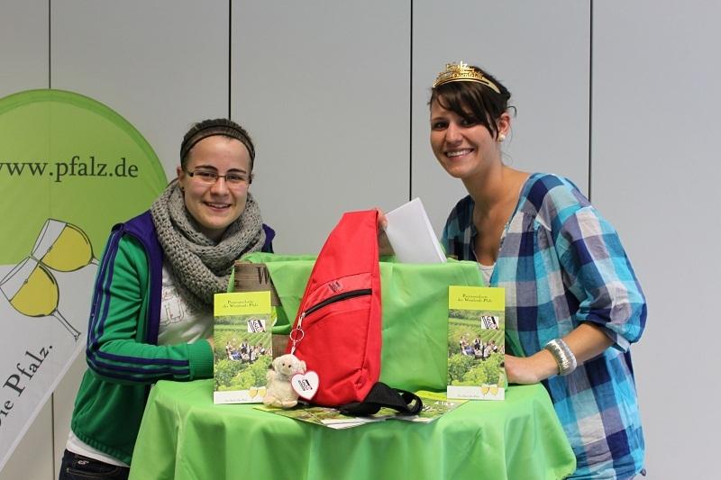 Zwei Frauen stehen an einem Tisch mit grüner Tischdecke, einem roten Rucksack und Informationsmaterialien über die Pfalz.