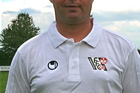 Portr&auml;t eines Mannes in einem wei&szlig;en Poloshirt mit Vereinslogo, stehend vor einem Fu&szlig;ballfeld unter bew&ouml;lktem Himmel.