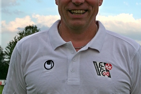 L&auml;chelnder Mann in einem wei&szlig;en Polo-Shirt mit Vereinslogos steht auf einem Fu&szlig;ballfeld unter einem bew&ouml;lkten Himmel.