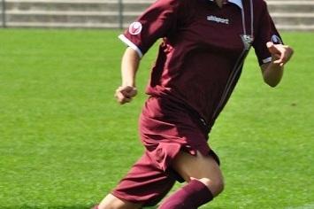 Frau in dunkelrotem Fu&szlig;balltrikot sprintet &uuml;ber den Rasen eines Fu&szlig;ballfeldes. Zuschauer im Hintergrund.