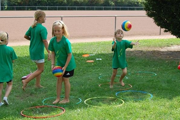 Kinder in gr&uuml;nen T-Shirts spielen im Freien mit bunten B&auml;llen und Hula-Hoops auf einer Wiese.