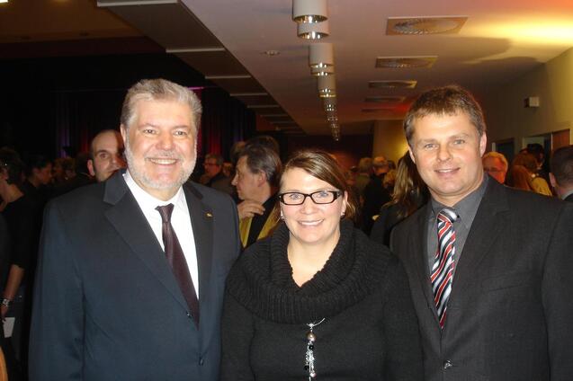 Drei Personen in formeller Kleidung stehen in einer Menschenmenge bei einer Veranstaltung im Hintergrund.