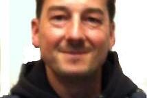 Portr&auml;t eines Mannes mit dunklem Haar, der in einem sportlichen Outfit mit Kapuze in die Kamera l&auml;chelt.