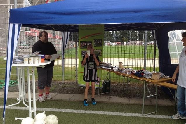 Junge in schwarz-wei&szlig;em Trikot h&auml;lt Urkunde am Zeltes, w&auml;hrend Erwachsene am Tisch mit Utensilien stehen.