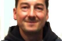 Portr&auml;t eines Mannes mit dunklem Haar, der in einem sportlichen Outfit mit Kapuze in die Kamera l&auml;chelt.