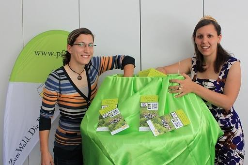 Zwei Frauen stehen an einem Tisch mit gr&uuml;ner Tischdecke, umgeben von Flyern, vor einem Banner im Hintergrund.