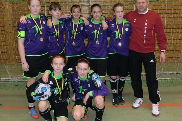 Sieben M&auml;dchen in lila Fu&szlig;balltrikots mit Medaillen stehen vor einer Torwand, begleitet von einem Trainer in rotem Shirt.