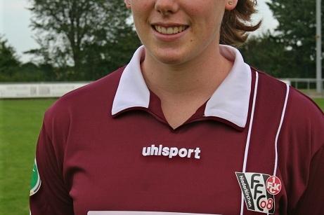 Portr&auml;t einer Fu&szlig;ballspielerin in einem dunkelroten Trikot mit Sponsorenlogo, im Hintergrund gr&uuml;ne Wiese und B&auml;ume.