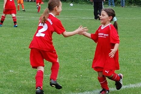 Zwei M&auml;dchen in roten Fu&szlig;balltrikots begr&uuml;&szlig;en sich auf einem gr&uuml;nen Rasenplatz nach einem Spielzug.