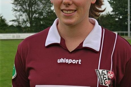 Portr&auml;t einer Fu&szlig;ballspielerin in einem dunkelroten Trikot mit Sponsorenlogo, im Hintergrund gr&uuml;ne Wiese und B&auml;ume.