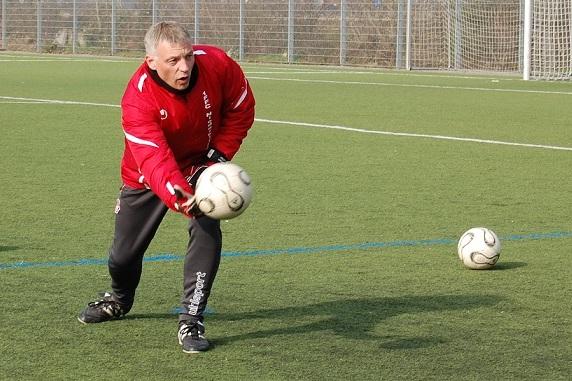 Trainer fängt einen Fußball auf einem Kunstrasenplatz, während ein weiterer Ball in der Nähe liegt.