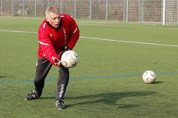 Trainer f&auml;ngt einen Fu&szlig;ball auf einem Kunstrasenplatz, w&auml;hrend ein weiterer Ball in der N&auml;he liegt.
