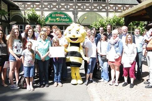 Gruppe von Menschen mit einem Bienenmaskottchen vor einem Gasthaus im Freien, alle l&auml;chelnd und zusammenstehend.