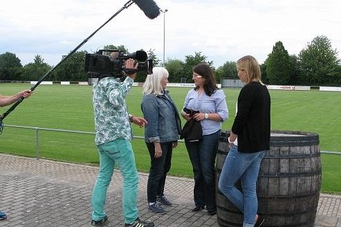 Drei Frauen stehen an einem Weinfass auf einem Fu&szlig;ballplatz, w&auml;hrend ein Kamerateam sie filmt.