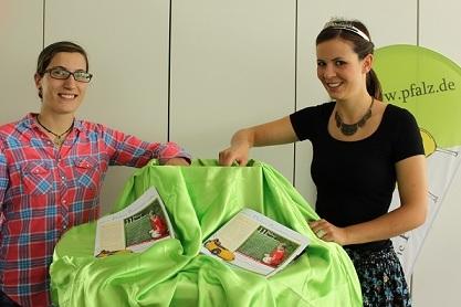 Zwei Frauen enth&uuml;llen eine gr&uuml;ne Tischdecke, darunter liegen Brosch&uuml;ren auf einem Tisch.
