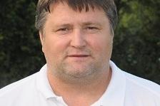 Portr&auml;t eines Mannes mit kurzem, dunklem Haar, der ein wei&szlig;es Polo-Shirt mit Logos tr&auml;gt, vor einer gr&uuml;nen Hintergrundlandschaft.