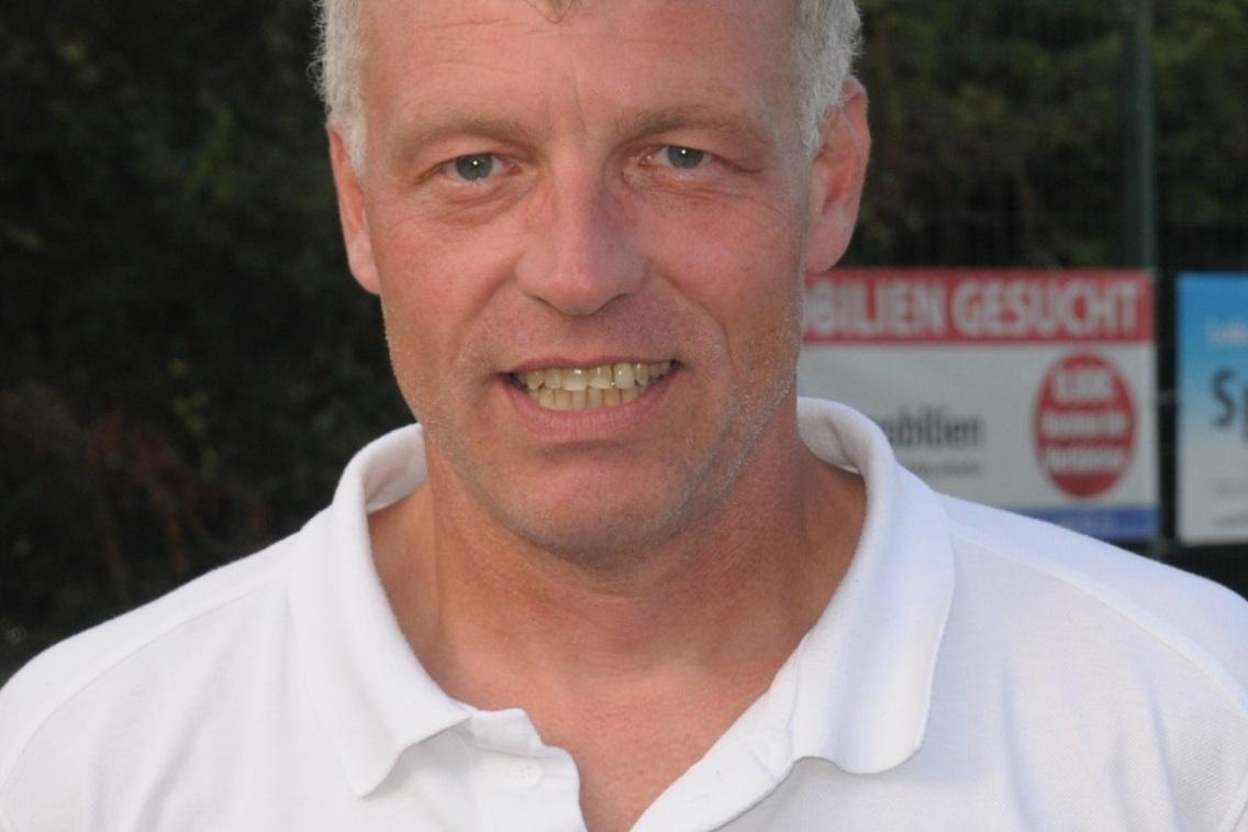 Portr&auml;t eines l&auml;chelnden Mannes mit kurzem, grauem Haar, in einem wei&szlig;en Polo-Shirt mit Vereinslogo.