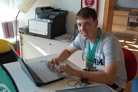 Junger Mann sitzt an einem Tisch mit Laptop, lächelt und trägt eine Medaille um den Hals. Im Hintergrund sind ein Drucker und Fenster.
