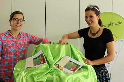 Zwei Frauen enth&uuml;llen eine gr&uuml;ne Tischdecke, darunter liegen Brosch&uuml;ren auf einem Tisch.