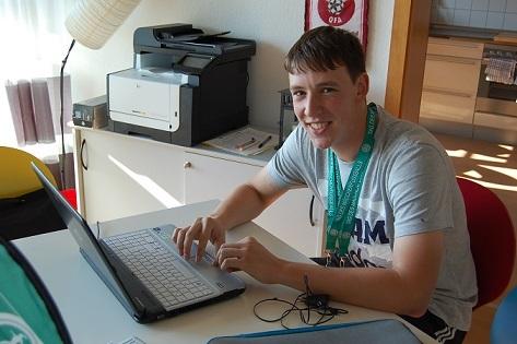 Junger Mann sitzt an einem Tisch mit Laptop, l&auml;chelt und tr&auml;gt eine Medaille um den Hals. Im Hintergrund sind ein Drucker und Fenster.