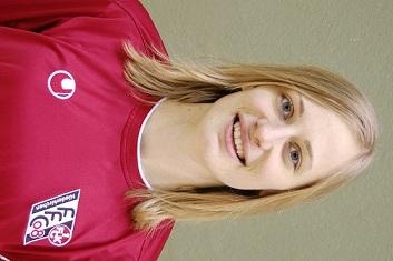 Portr&auml;t einer l&auml;chelnden Frau in rotem Sporttrikot mit Vereinslogo, vor einer gr&uuml;nen Wand.