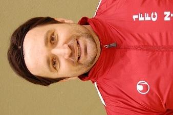 Portr&auml;t eines Mannes mit braunen Haaren, der in einem roten Sportjackett grinst, vor einer gr&uuml;nen Wand.