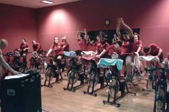 Gruppe von Personen in Rot auf Spinning-Bikes in einem Fitnessraum mit Handt&uuml;chern auf den Ger&auml;ten.