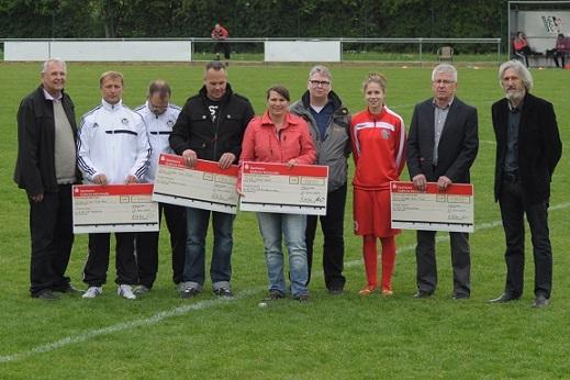 Gruppenfoto von acht Personen auf einem Fu&szlig;ballfeld, die Schecks f&uuml;r einen finanziellen Zuschuss halten.