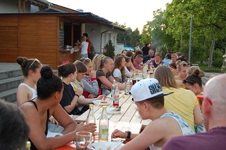 Gruppe von Jugendlichen an einem langen Tisch im Freien, essen und trinken bei Sonnenuntergang.