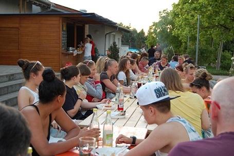 Gruppe von Jugendlichen an einem langen Tisch im Freien, essen und trinken bei Sonnenuntergang.