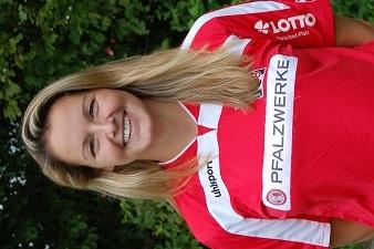 Portr&auml;t einer l&auml;chelnden Frau in rotem Fu&szlig;balltrikot mit Sponsorenlogos, Hintergrund mit B&auml;umen.