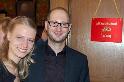 Paar posiert l&auml;chelnd vor einer T&uuml;r mit rotem &bdquo;Bitte nicht st&ouml;ren! Trauung&ldquo;-Schild.