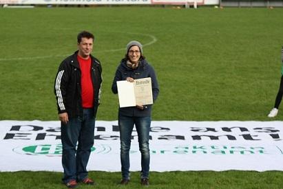 Zwei Personen stehen auf einem Fu&szlig;ballfeld und halten eine Urkunde vor einem gro&szlig;en Banner mit der Aufschrift &bdquo;Danke ans Ehrenamt&ldquo;.