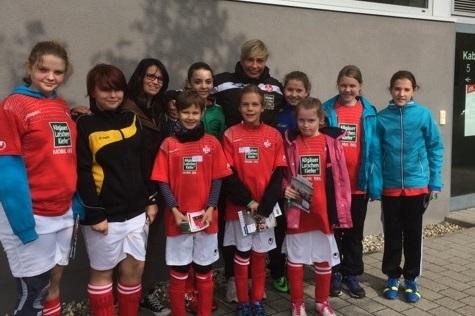 Gruppe von M&auml;dchen in Fu&szlig;balltrikots mit Trainerin vor einem Geb&auml;ude, alle l&auml;chelnd und mit Informationsbl&auml;ttern in der Hand.