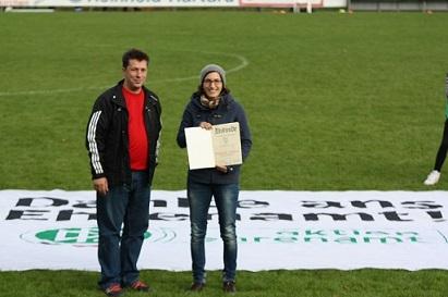 Zwei Personen stehen auf einem Fu&szlig;ballfeld und halten eine Urkunde vor einem gro&szlig;en Banner mit der Aufschrift &bdquo;Danke ans Ehrenamt&ldquo;.