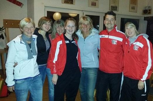 Gruppe von sieben Personen in Sportbekleidung, l&auml;cheln gemeinsam in einem Innenraum mit dekorativen Elementen.