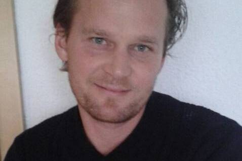 Portr&auml;t eines Mannes mit kurzem, unordentlichem Haar, der in die Kamera l&auml;chelt, vor einem hellen Hintergrund.