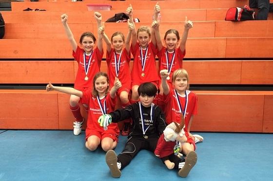 Gruppe von sieben Kindern in roten Trikots mit Medaillen, die jubelnd auf einer Tribüne stehen.