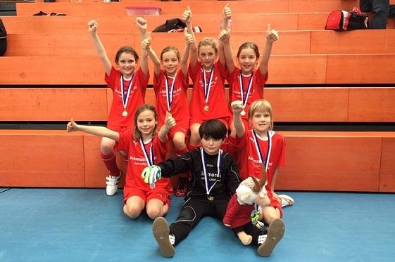 Gruppe von sieben Kindern in roten Trikots mit Medaillen, die jubelnd auf einer Trib&uuml;ne stehen.